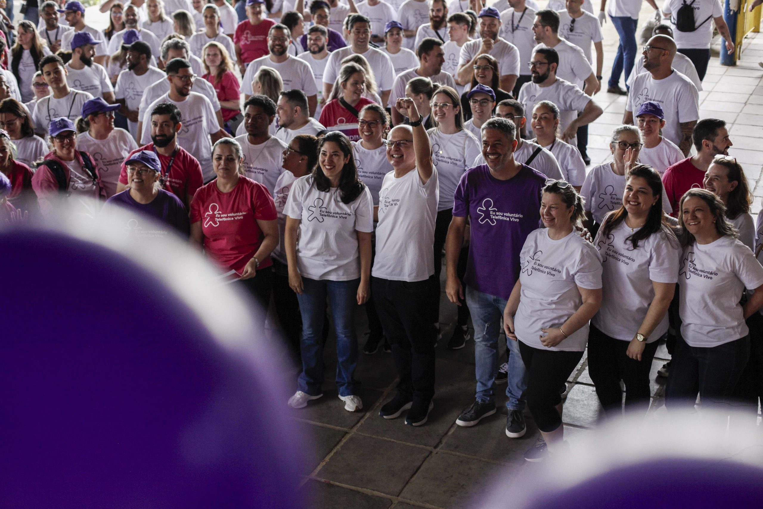 Multidão de voluntários se reúne no pátio da escola, em frente ao palco.