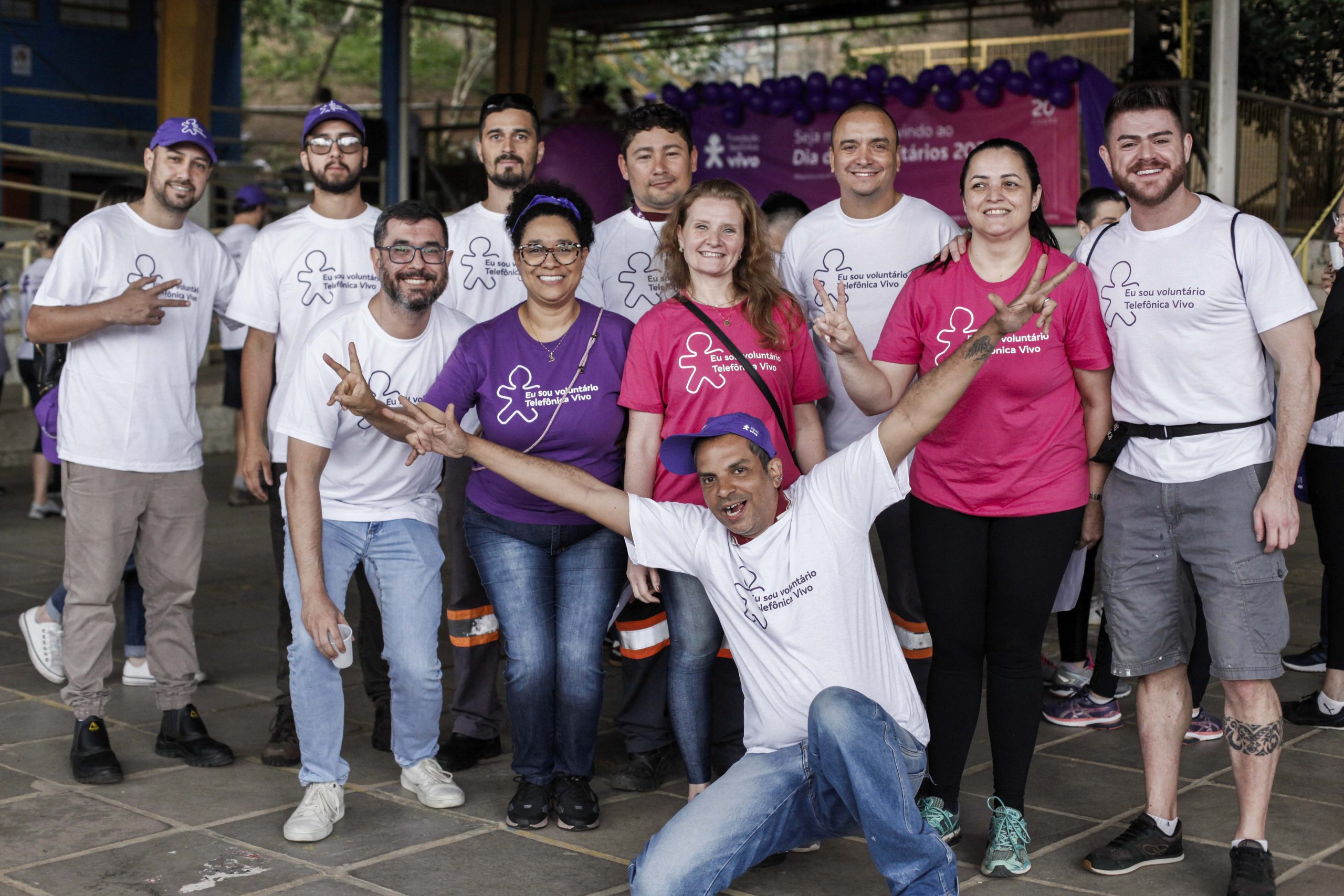 Grupo de voluntários posa para foto, dispostos lado a lado e em duas filas, com um homem à frente agachado e com os braços abertos, fazendo sinal de vitória com as mãos.