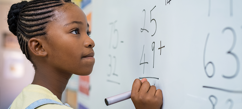 A imagem mostra uma aluna negra jovem em frente a uma lousa, onde há uma conta simples de matemática.