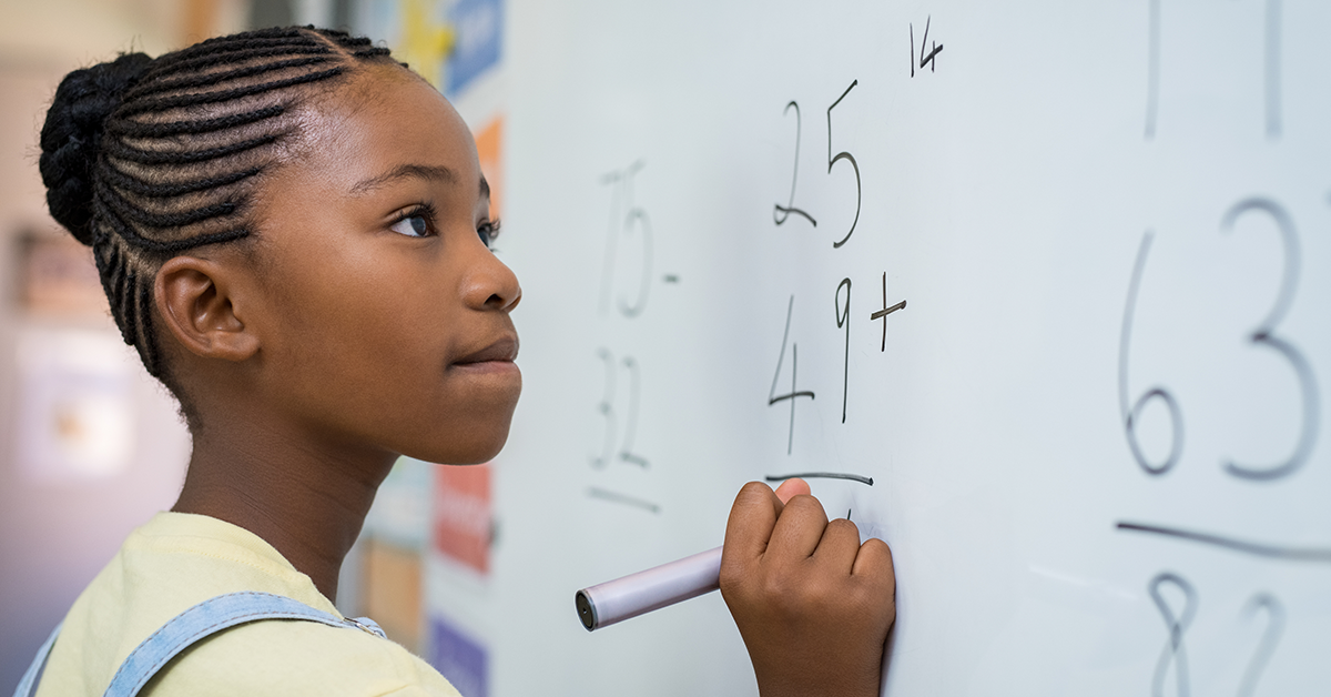 A imagem mostra uma aluna negra jovem em frente a uma lousa, onde há uma conta simples de matemática.