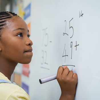 A imagem mostra uma aluna negra jovem em frente a uma lousa, onde há uma conta simples de matemática.