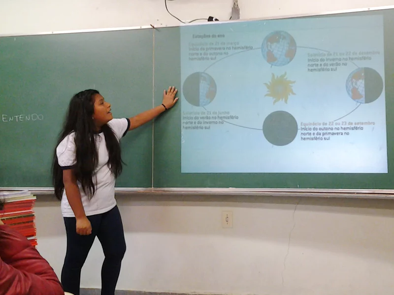 Menina de traços bolivianos com a mão apoiada na lousa olha para projeção enquanto faz apresetnação na sala de aula