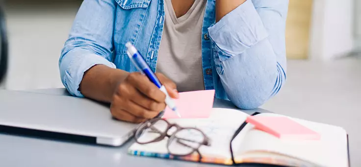 Na imagem é possível ver a silhueta de uma pessoa escrevendo termos sobre educação em um pedaço de papel. Há um par de óculos sobre a mesa