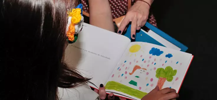 Criança está lendo página desenhada de livro publicado pelo Estante Mágica, projeto que publica histórias escritas por crianças.