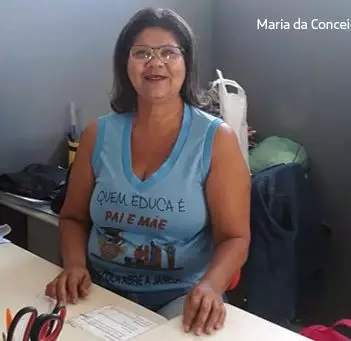 Gestora Conceição sorri para a câmera sentada em sua mesa. Ela usa blusa azul onde se lê: “Quem educa é pai e mãe”.