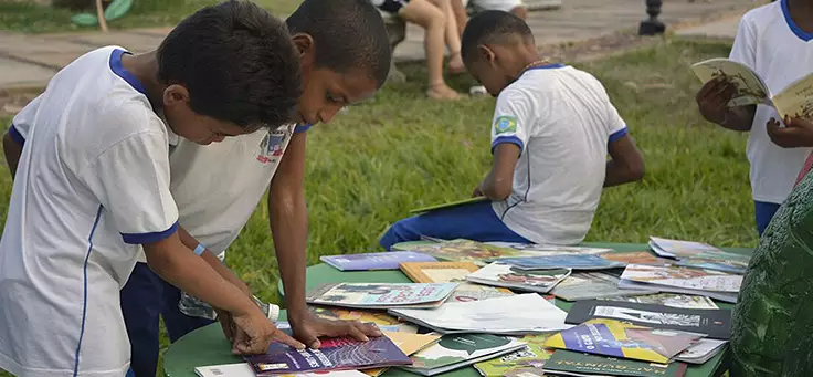 Crianças de uniforme escolar mexem em livros expostos em uma mesa ao ar livre
