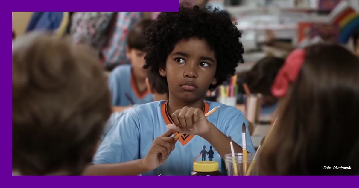 Imagem mostra um menino negro, com uniforme escolar, sentado em uma mesa com outras crianças. Ele segura um lápis de cor. Sobre a mesa, há outros lápis, pincéis, papel e um frasco de tinta