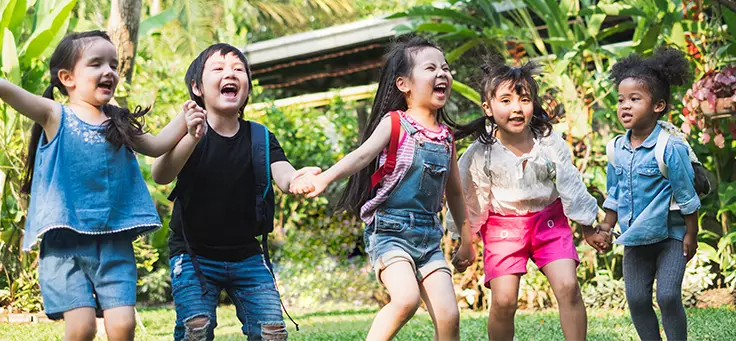 Imagem mostra um grupo de crianças de mãos dadas sorrindo ao ar livre com uma vegetação ao fundo