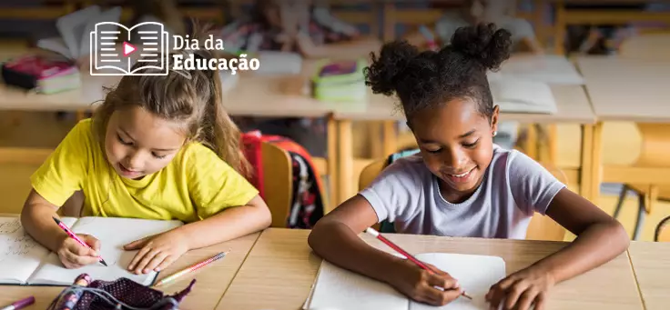 Imagem mostra duas meninas em destaque dentro de uma sala de aula. Elas estão sentadas uma ao lado da outra, escrevendo em cadernos.