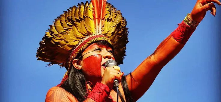 Célia Xakriabá é uma das lideranças indígenas que se destacam no Brasil. Ela está usando um cocar amarelo e laranja, além de pintura corporal enquanto fala ao microfone e gesticula com o braço esquerdo levantado.