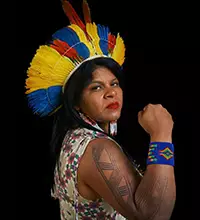 Sônia Guajajara é uma das lideranças indígenas que se destacam no Brasil. Ela está posando de lado, usando um cocar amarelo e azul, além de pintura corporal e adereço no braço direito erguido em pose. com o braço esquerdo levantado.