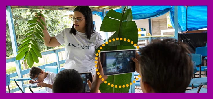 A foto mostra uma formadora do Projeto Aula Digital, que usa uma camiseta com o nome do programa, mostrando uma planta que está em suas mãos para um grupo de alunos. Um deles, segura um tablet com a câmera apontada na direção dela.