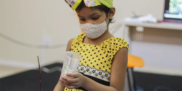 Menina usando máscara segura copo com muda de feijão em atividade na Associação Colorindo a Vida durante a Semana dos Voluntários.