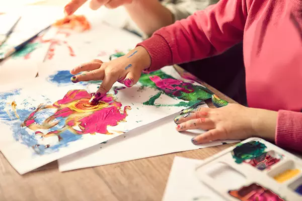 A imagem mostra as mãos de uma criança em destaque usando tinta para pintar com os dedos em uma folha de papel
