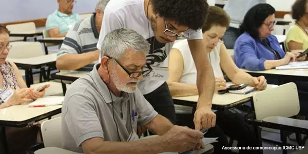 Aluno voluntário explica para uma pessoa idosa o funcionamento de um celular em sala de aula