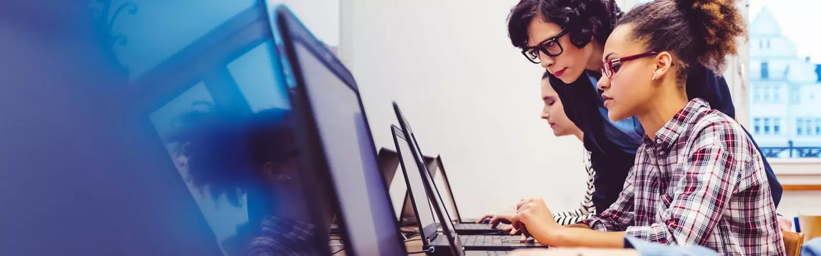 Imagem mostra dois jovens em sala de aula utilizando computadores. Eles estão acompanhados por uma educadora que parece estar observando o que uma das jovens está fazendo.