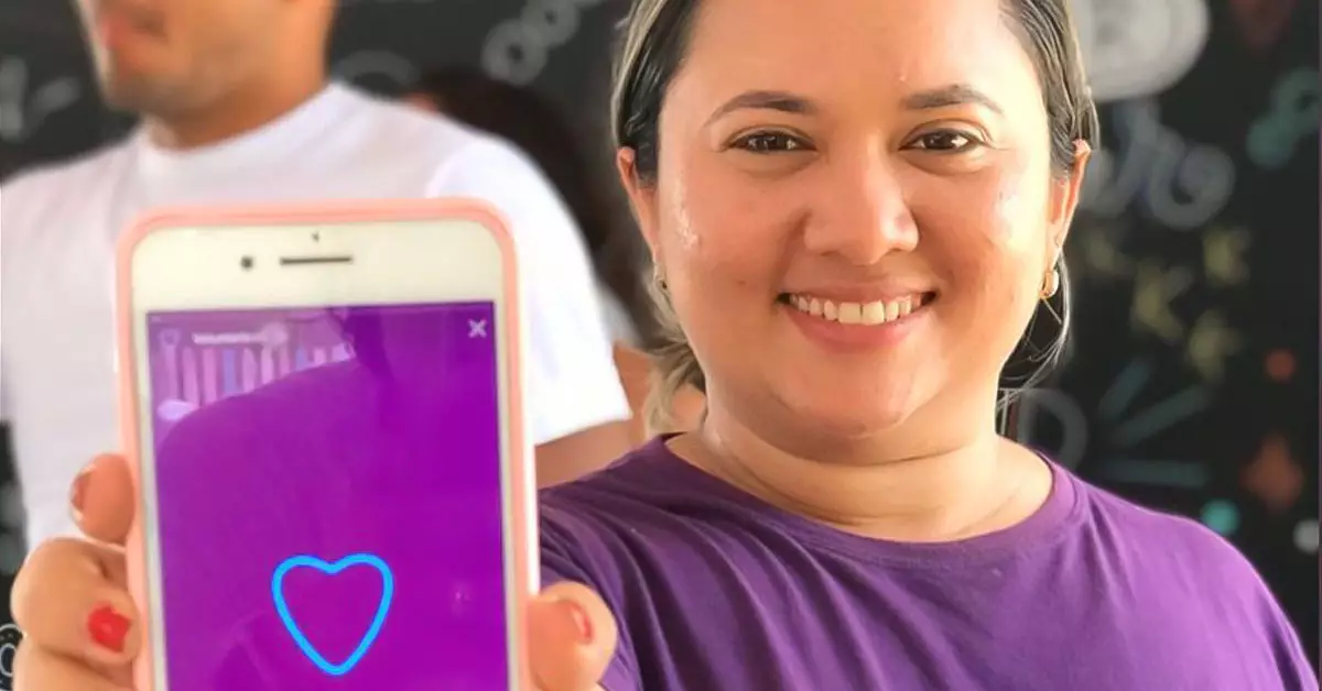 Imagem mostra a voluntária Geciclelma. Ela usa uma camiseta na cor roxa, está segurando um celular com a mão direita e mostrando ele para a câmera. Ela está sorrindo. Na tela do celular, há uma imagem na cor roxa com a ilustração de um coração.