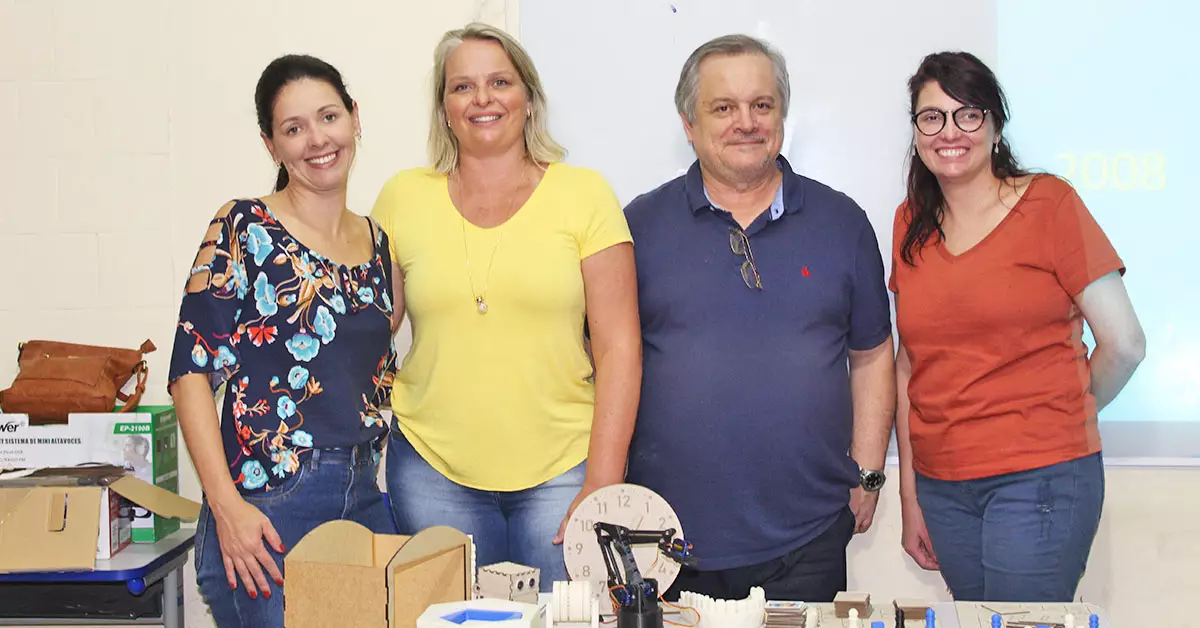 Imagem mostra as três professoras e um professor que fazem parte da equipe responsável pelo projeto vencedor do prêmio enlighted. Eles estão lado a lado em um ambiente que parece ser um laboratório. É possível ver uma mesa e alguns objetos pedagógicos em cima dela.