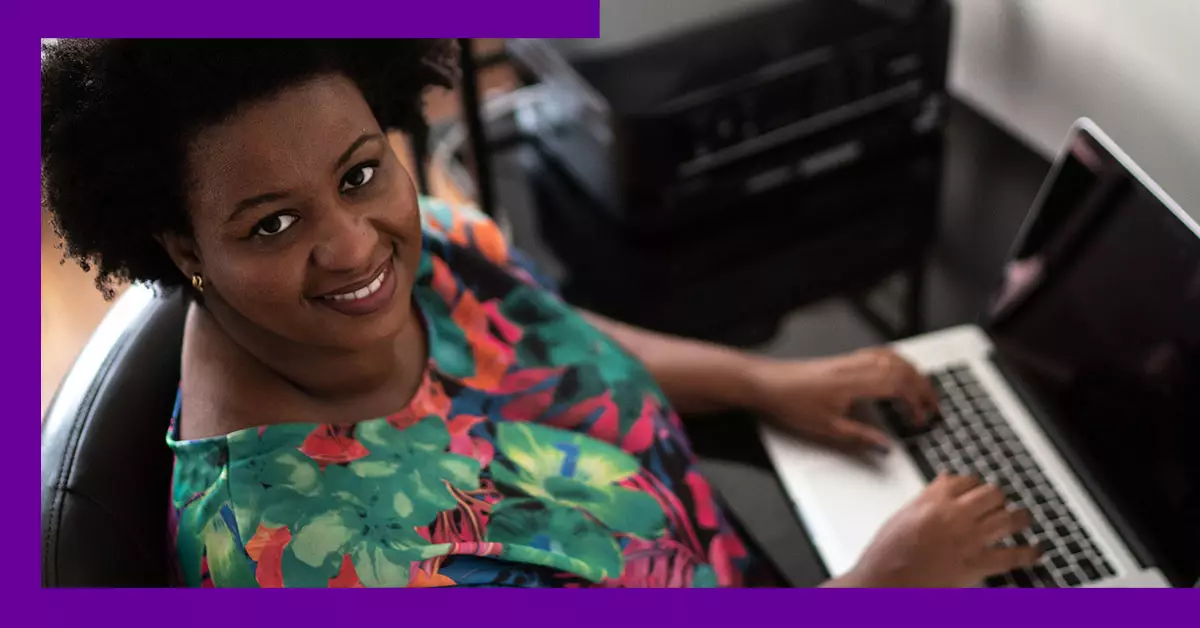 Professora usa um laptop e sorri para a foto. Ela é uma mulher negra e usa uma blusa verde com estampa de flores