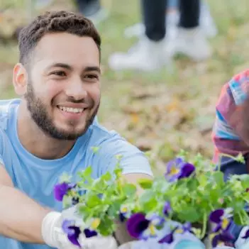 Imagem mostra três jovens em um local que parece ser um parque. Um dos jovens está segurando um vaso de flores e entregando para uma das jovens. A terceira jovem observa os dois jovens interagindo.