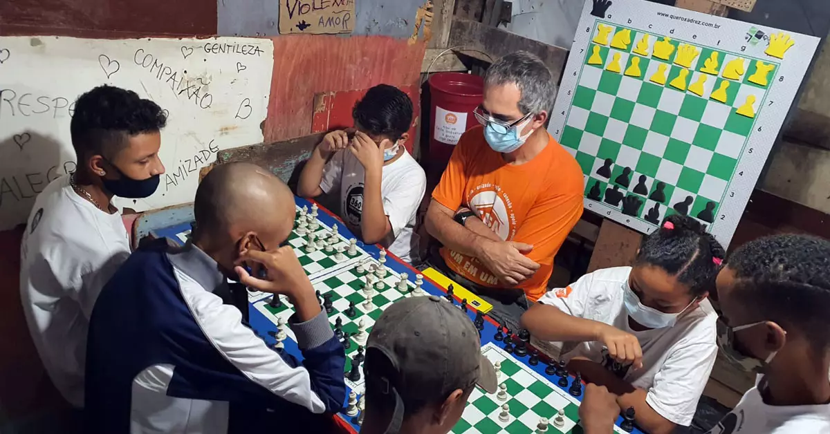 Imagem mostra alunos do projeto Casa Xadrez em volta de uma mesa onde há três tabuleiros do jogo. Eles estão acompanhados de um adulto, que parece ser o professor, e estão concentrados observando as peças do jogo