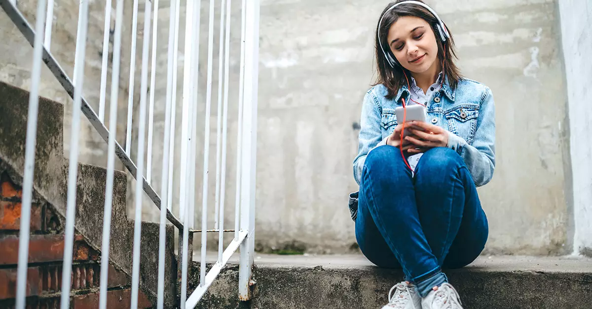 imagem mostra uma jovem sentada em uma escada segurando um celular nas mãos e fones de ouvido.