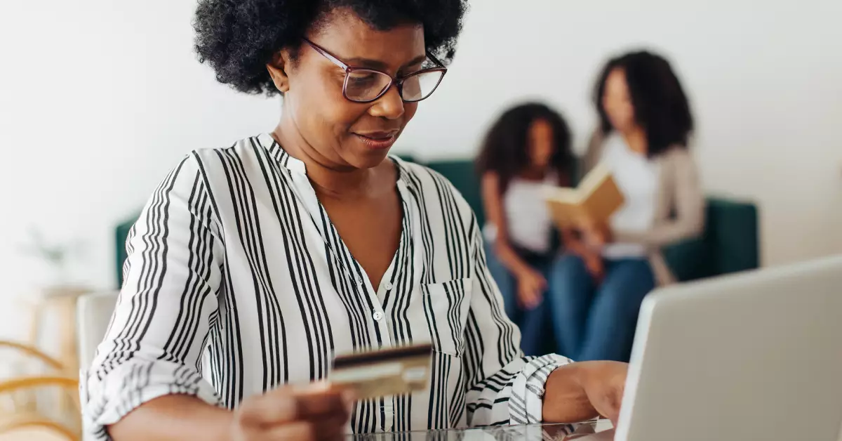 Imagem mostra uma mulher negra utilizando um notebook. Ela segura um cartão de crédito na mão direita, o que dá a entender que ela está fazendo uma transação financeira pela internet. Ao fundo, é possível ver uma mulher e uma menina sentadas em um sofá.