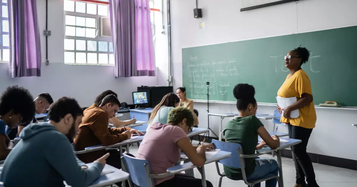 Sala de aula com professor de pé, alunos sentados na carteira fazendo anotações e lousa verde.