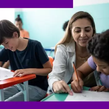 Imagem mostra uma professora auxiliando dois alunos em sala de aula. A professora é branca e tem cabelos longos. O aluno é branco e a aluna é negra.