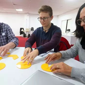 Imagem mostra três professores participando de uma dinâmica, são dois homens e uma mulher. Eles estão sentados em volta de uma mesa, mexendo com objetos circulares amarelos que estão sobre a mesa.