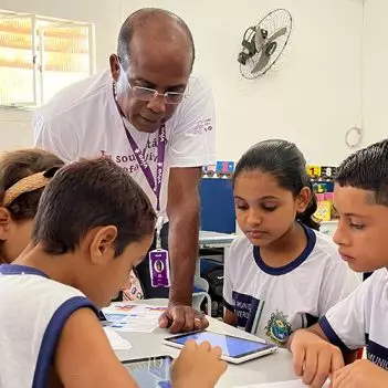 Imagem mostra um voluntário negro orientando quatro alunos em uma sala de aula. O voluntário usa óculos. Os alunos estão sentados e observam tablets que estão em cima da mesa.