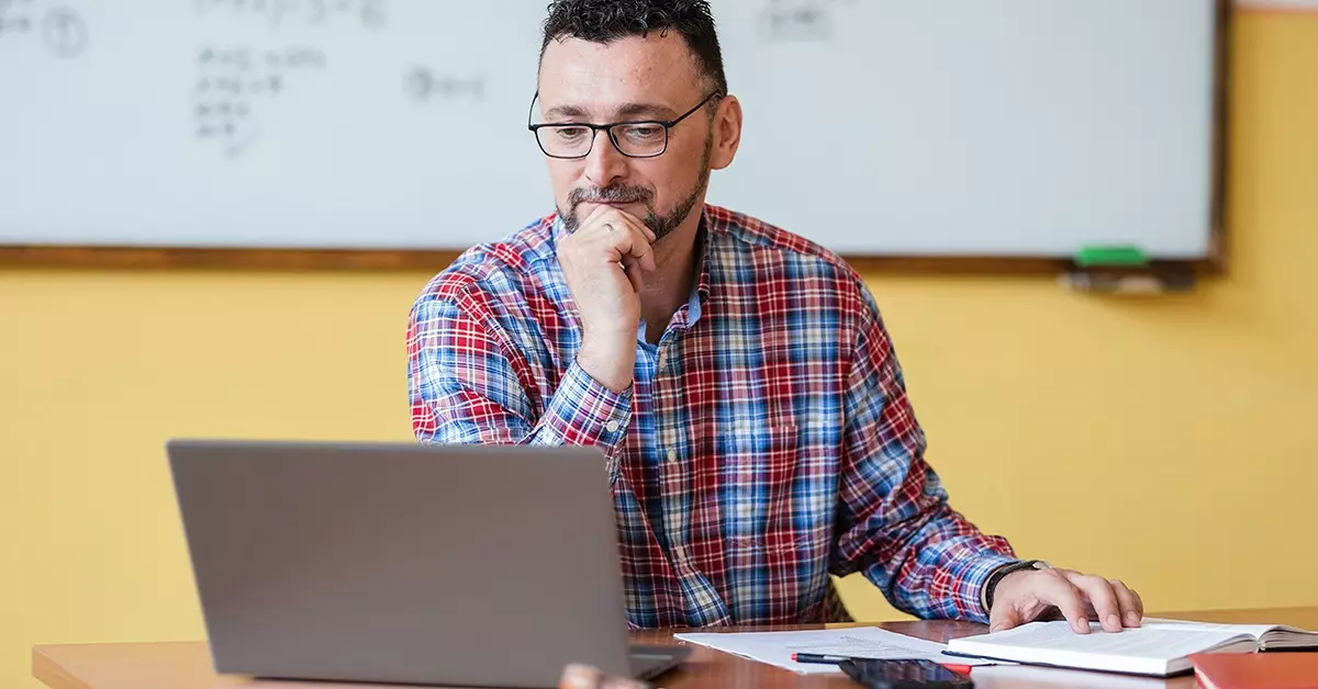 Imagem mostra um professor manuseando um notebook e um mouse. Ele está em uma sala de aula.