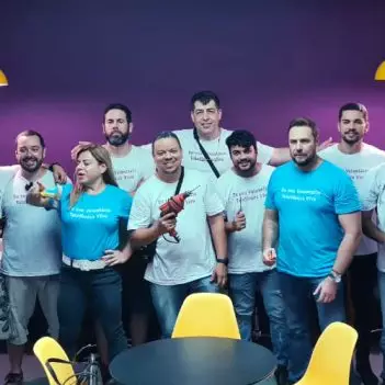 Imagem mostra um grupo de 12 voluntários; são 10 homens e 2 mulheres. Eles estão reunidos em uma sala, ao fundo há uma parede na cor roxa. Eles usam camisetas com a frase sou voluntário telefônica