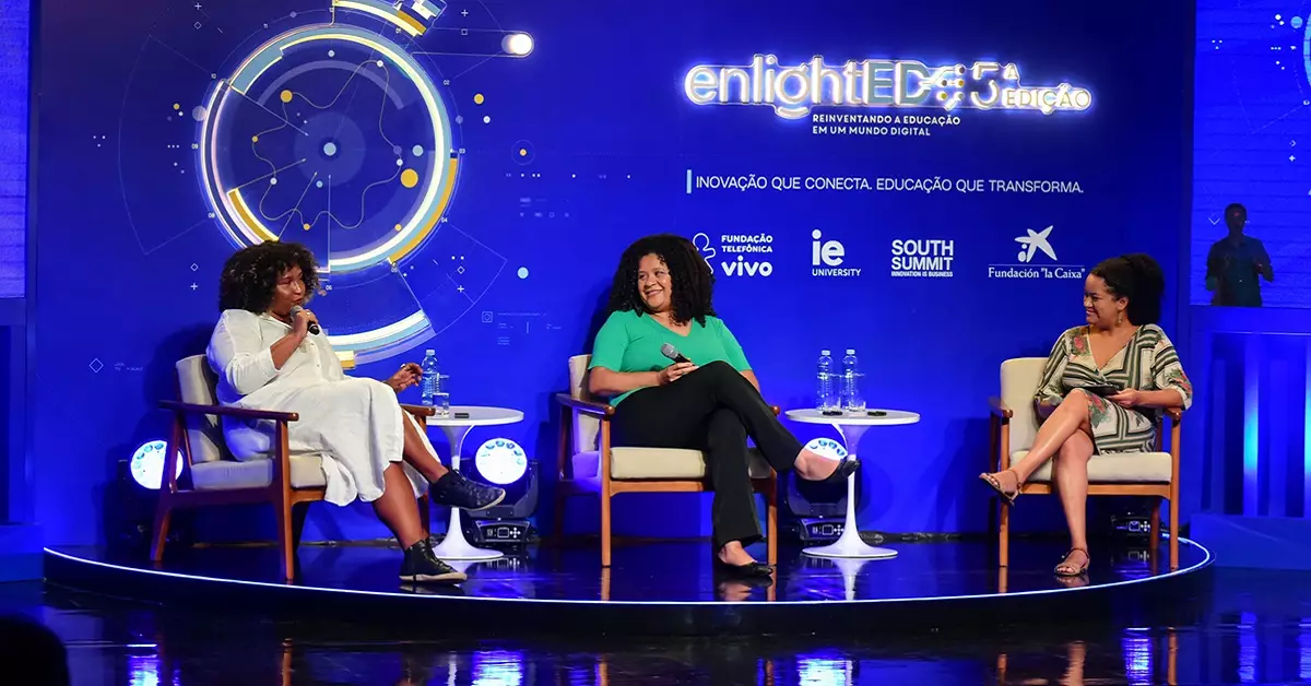 Imagem mostra o palco do enlighted com três palestrantes no palco. Elas são mulheres negras, estão sentadas, uma delas está com o microfone falando ao público