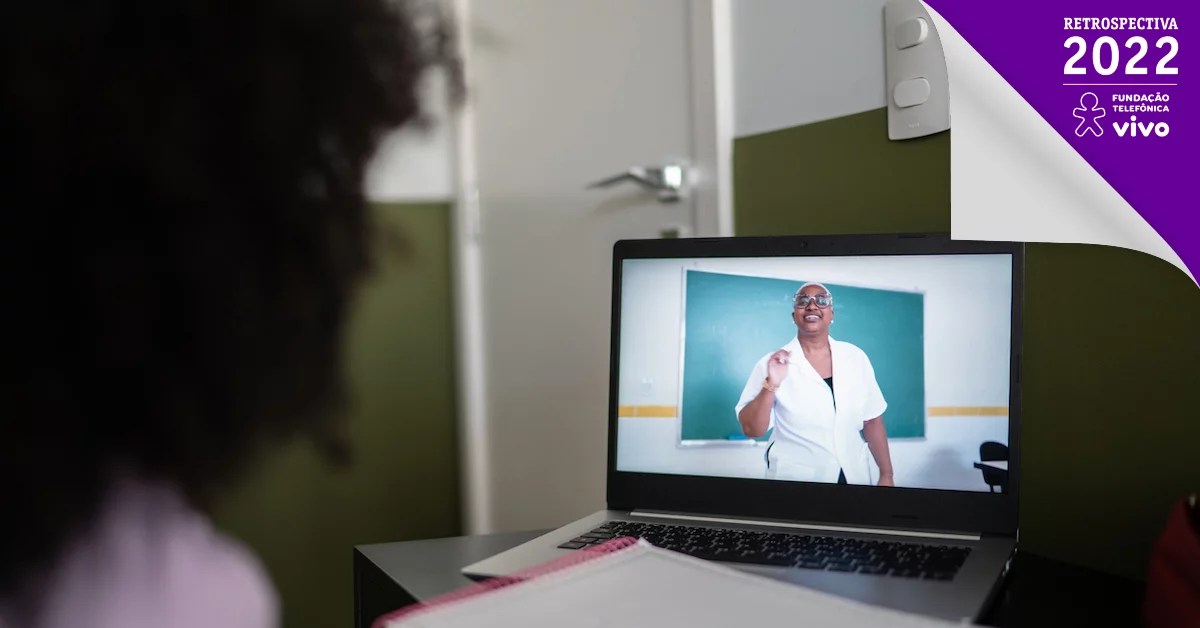 De costas, menina olha para a tela de um computador, onde uma professora dá aula em frente à lousa.