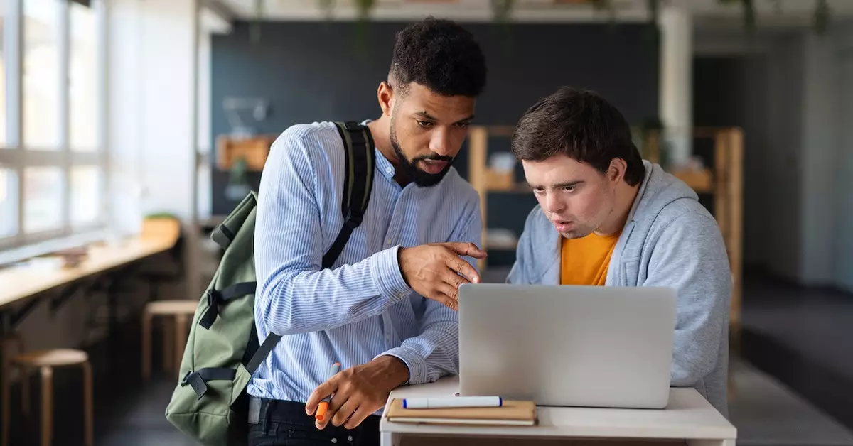 Em uma sala de aula, um estudante negro conversa com um estudante com síndrome de down, em frente a um laptop aberto