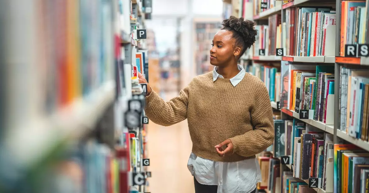 Jovem mulher negra veste camisa azul clara e suéter bege. Ela está na biblioteca e escolhe livros sobre ciência de dados