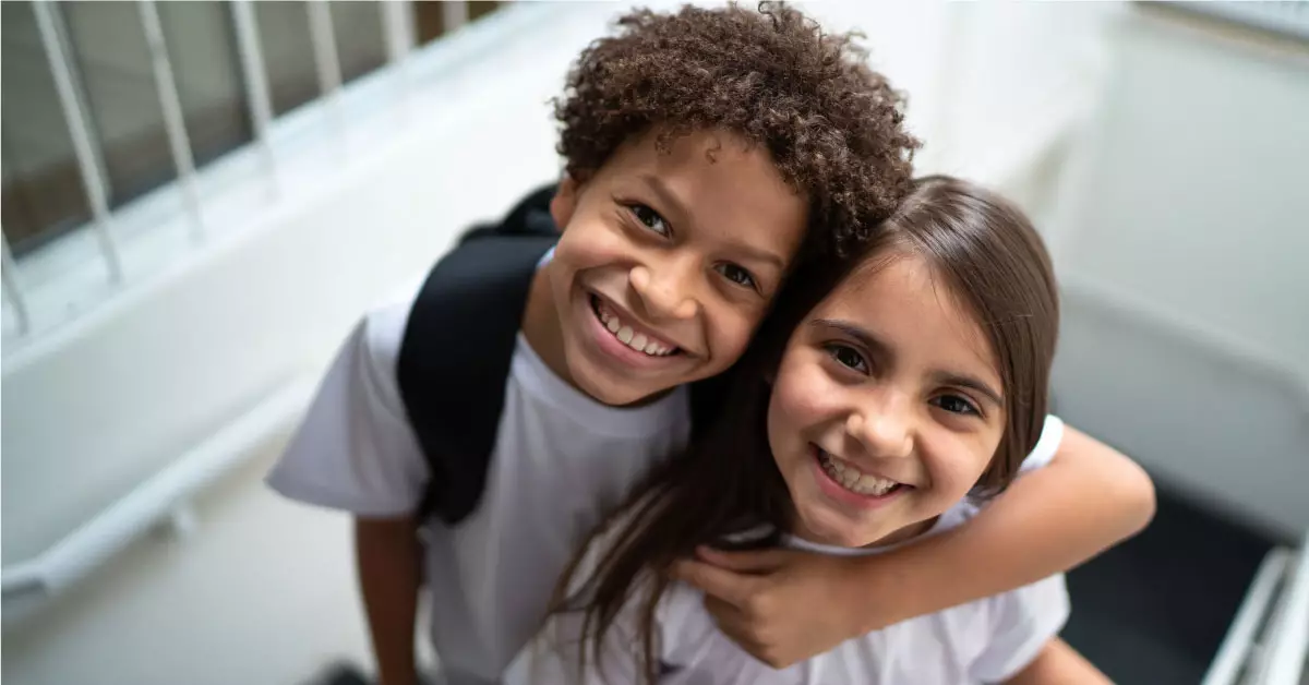 Imagem mostra duas crianças de ensino fundamental 1. O menino está à esquerda da imagem e abraça a menina que está à direita. Os dois usam camiseta branca. O menino está de mochila preta nas costas.