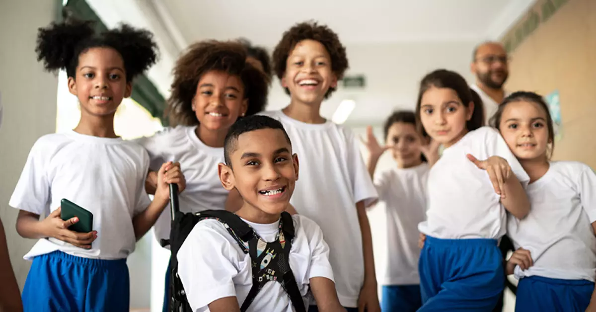 A imagem mostra um grupo de sete crianças da educação infantil. Todos vestem uniforme, com camiseta branca e calça azul royal. Em primeiro plano, está um menino na cadeira de rodas. Os demais estudantes estão sorrindo e em pé em volta dele. Ao fundo do grupo, está a cabeça desfocada de um professor.