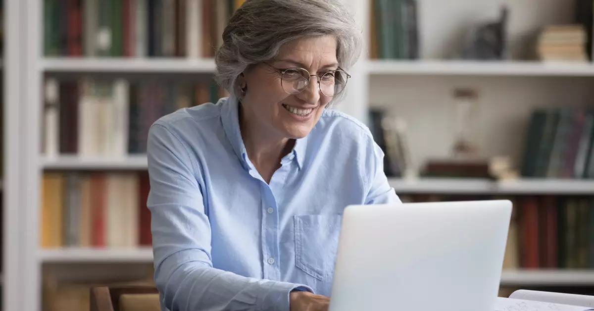 A foto mostra uma mulher sorrindo enquanto olha para um notebook branco. Ela tem cabelos grisalhos e usa óculos de aro redondo. Está vestindo uma camisa de manga longa azul clara. Atrás dela, está uma estante com livros.