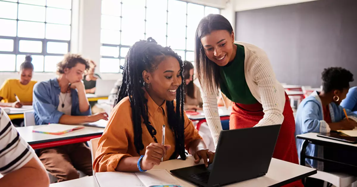 Imagem mostra uma sala de aula. Em primeiro plano,uma estudante negra está usando um notebook e uma professora negra está a auxiliando. Ao fundo, podemos ver outros estudantes sentados em suas mesas