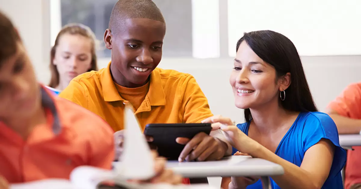 Imagem mostra um aluno negro sendo orientado por sua professora, branca, no uso de um tablet