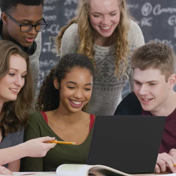 A imagem mostra um grupo de seis estudantes em uma sala de aula. Quatro deles, dois meninos e duas meninas, estão sentados à mesa olhando para um notebook preto. Há também um menino e uma menina em pé, debruçado no mesa e olhando para a tela do notebook. Atrás há uma lousa com alguns conteúdos escritos. E também mapas pendurados na parede.