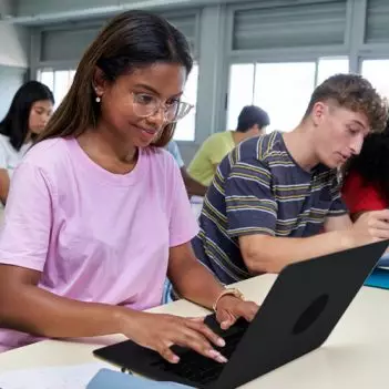 A imagem mostra um grupo diverso de estudantes adolescentes. Eles estão sentados em duas bancadas de estudos com cadernos, canetas e notebooks. Em primeiro plano, a imagem mostra quatro estudantes: duas meninas e dois meninos. Em segundo plano, é possível ver mais quatro estudantes, também dois meninos e duas meninas.