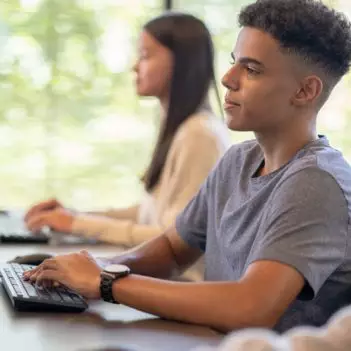 A imagem mostra três adolescentes sentados em frente a computadores digitando. Em primeiro plano, está um menino pardo que veste camiseta cinza e usa relógio no punho. Em segundo plano, desfocadas, estão uma menina loira com blusa branca de manga comprida e uma menina oriental de blusa de lã branca.