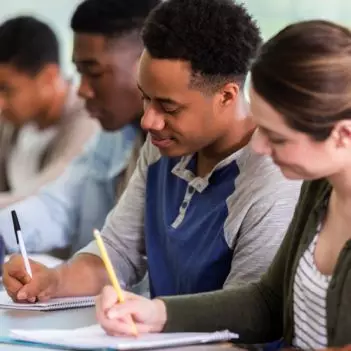 Imagem mostra grupo de estudantes em sala de aula, sentados, com canetas e lápis em mãos, escrevendo em cadernos. Vemos estudantes homens negros e uma estudante mulher branca