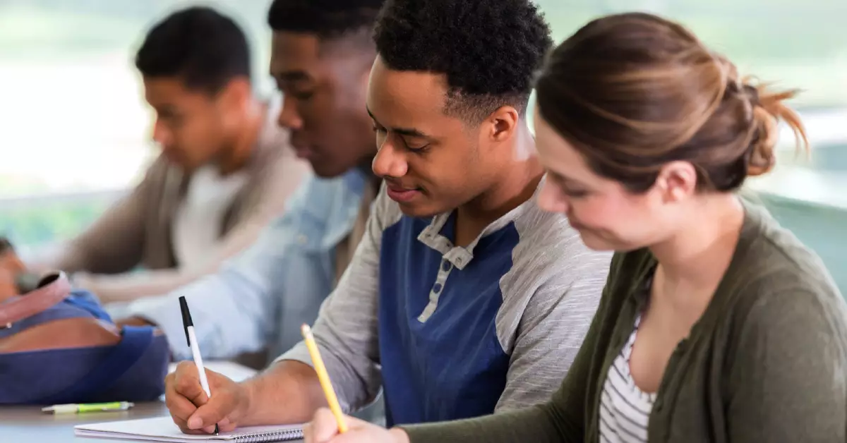 Imagem mostra grupo de estudantes em sala de aula, sentados, com canetas e lápis em mãos, escrevendo em cadernos. Vemos estudantes homens negros e uma estudante mulher branca