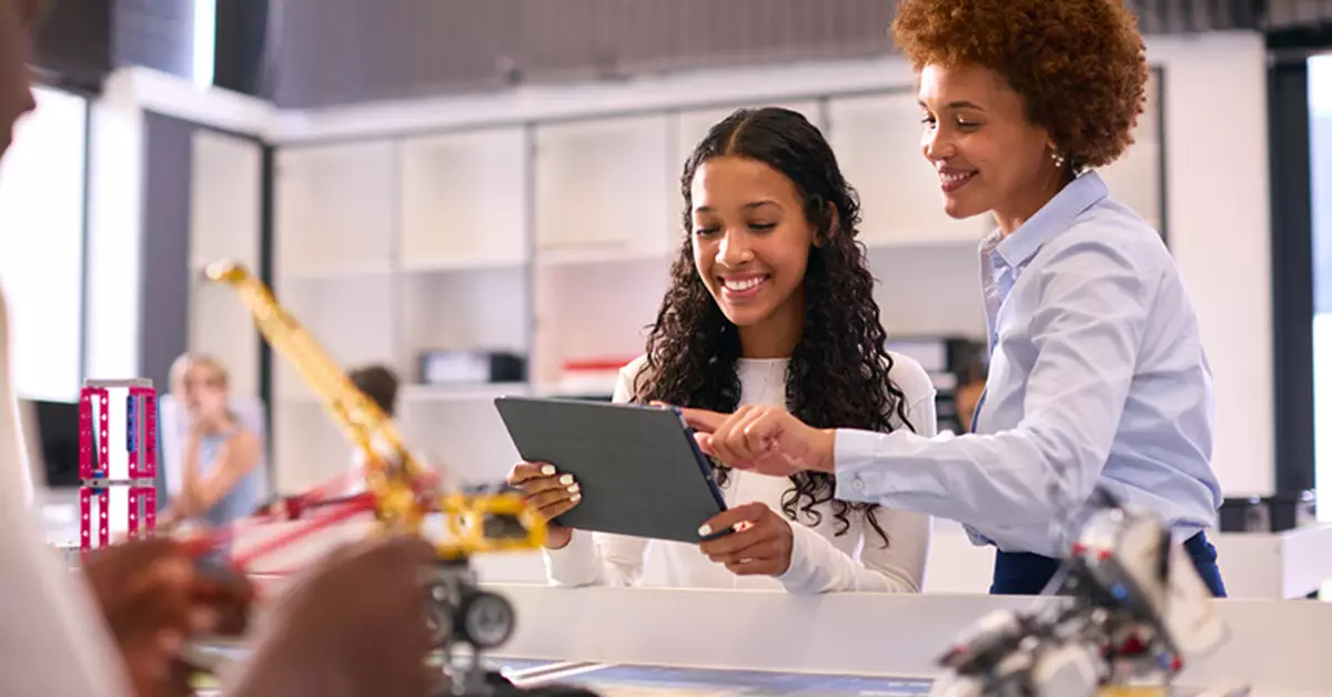 A imagem mostra uma professora e uma estudante interagindo com um tablet. A estudante é negra, tem cabelos longos e cacheados e está vestindo uma blusa branca de manga comprida. A professora é negra, tem um cabelo black power e está usando uma camisa azul clara e calça azul marinho. As duas estão em pé e em frente a uma bancada branca que contém alguns itens de robótica