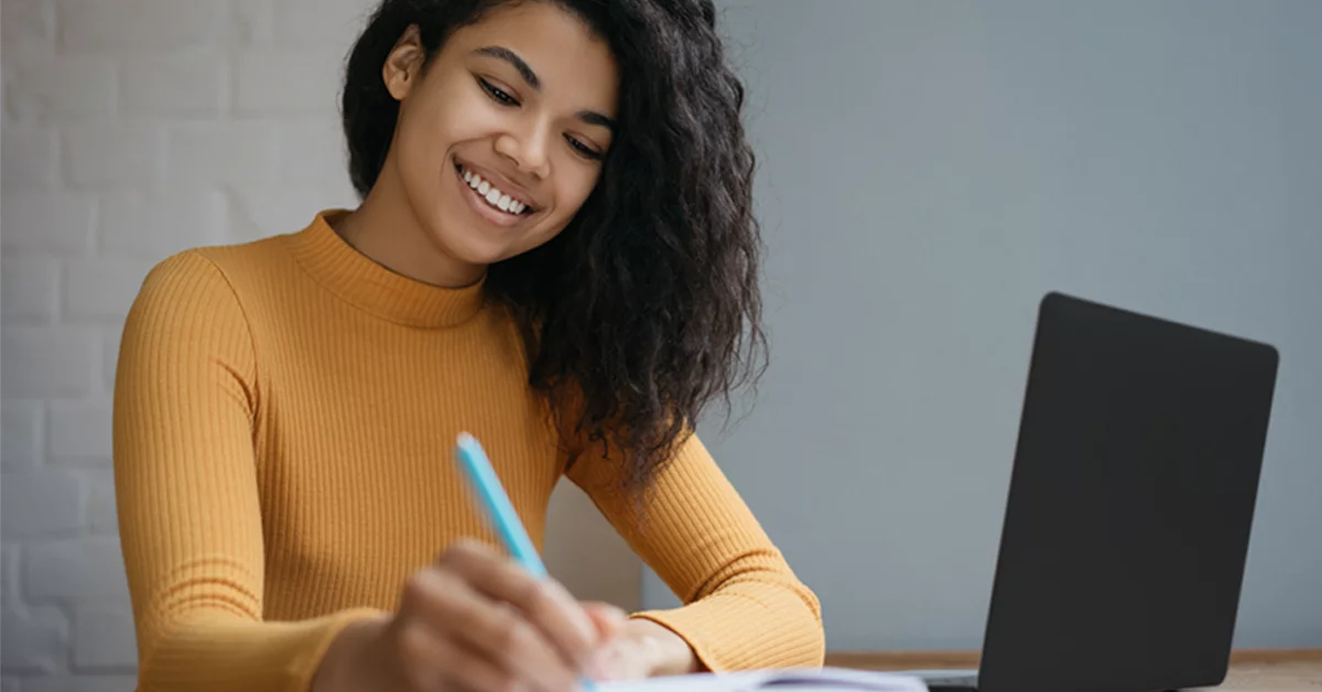 Imagem mostra uma estudante negra, de cabelos longos, utilizando um notebook que está sobre uma mesa, e com uma caneta na mão direita